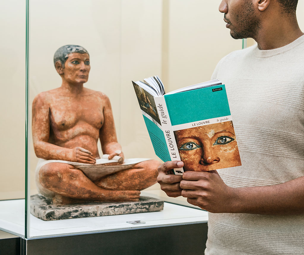 Visiteur tenant le nouveau guide du Louvre devant le Scribe accroupi